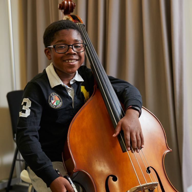 A boy with black hair and wearing a black long sleeved top sits with his violin. He has a big smile across his face.